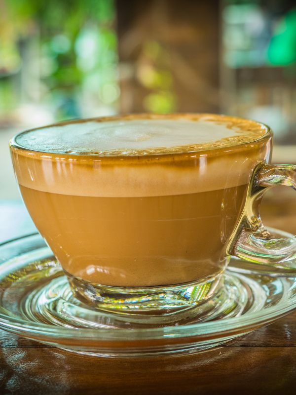 Cappuccino in a clear glass cup with a thick layer of milk foam on top, served on a saucer.