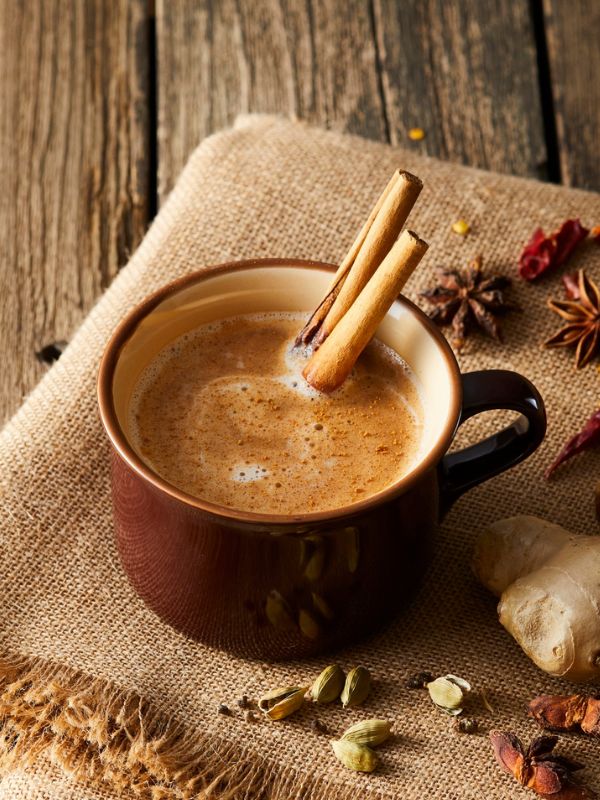 Hot chai latte in a brown mug with cinnamon sticks, surrounded by spices on a rustic wooden table.